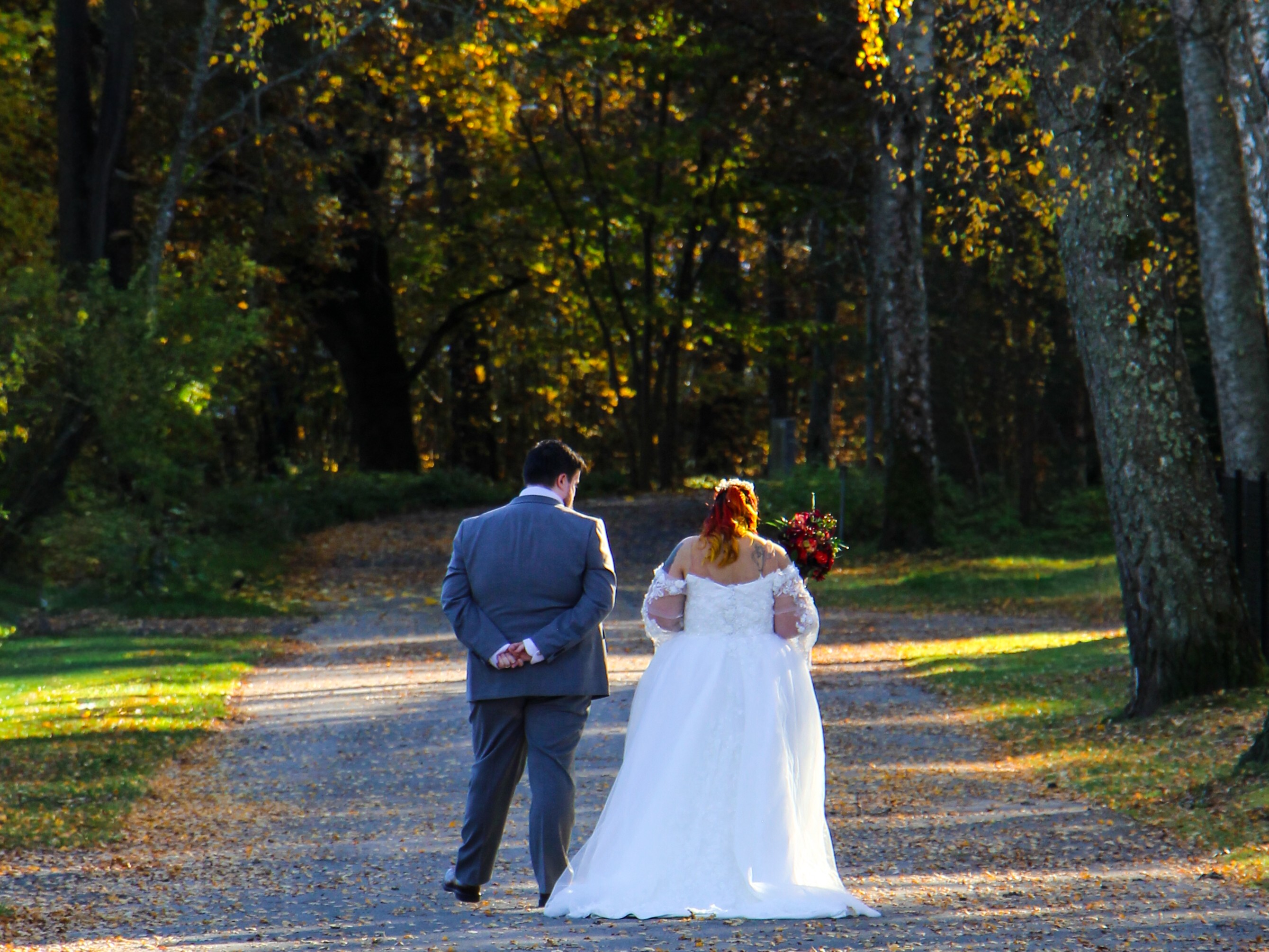 A bride and groom walk away from the camera down a tree-lined path in the fall. The bride wears a white wedding dress and the groom a grey suit.